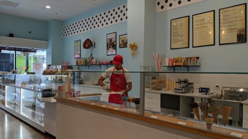 A worker in a red apron prepares ice cream at a colorful shop with glass displays and a menu on the wall.
