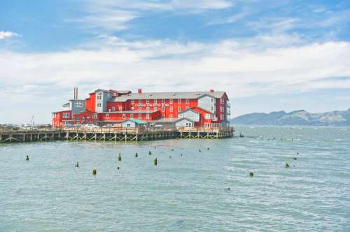 A red building on stilts by the water, with mountains in the background and a cloudy sky above.