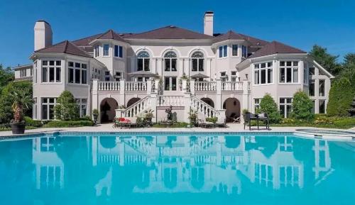 A luxurious mansion with large windows and a pool in the foreground, surrounded by lush greenery and a clear blue sky.