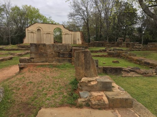 Ruins of an old structure surrounded by grassy areas and trees, with stone foundations and remnants visible.