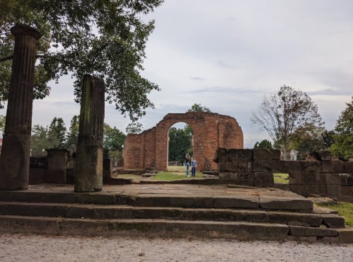 Two people stand among the ruins of an old brick structure, framed by trees and a cloudy sky.