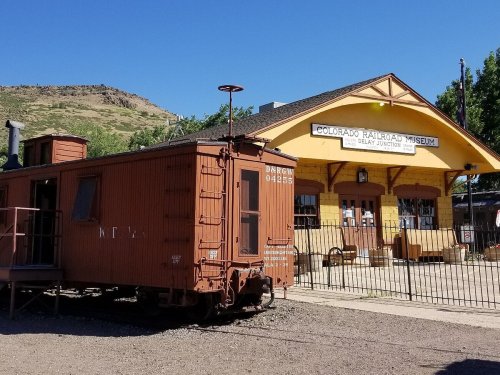 A vintage train car in front of the Colorado Railroad Museum building at Delay Junction, surrounded by greenery.