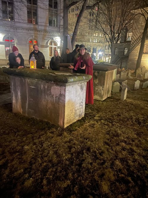A person in a red cloak stands by a stone grave, surrounded by a small group of people at night.