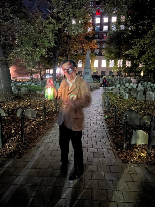 A person in a blazer stands in a dimly lit graveyard, holding colorful lights among gravestones and autumn foliage.