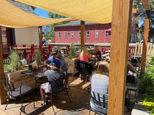 Outdoor dining area with people sitting at tables under a shaded canopy, enjoying meals on a sunny day.