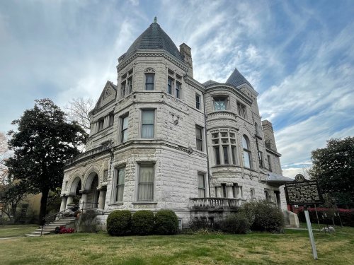 A large, historic stone mansion with turrets, surrounded by greenery and a cloudy sky.