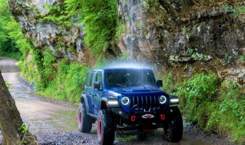A blue off-road Jeep driving along a scenic, rocky path surrounded by lush greenery.