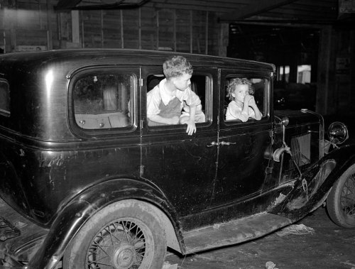 Two children sit in an old black car, one leaning out the window while the other looks thoughtfully inside.