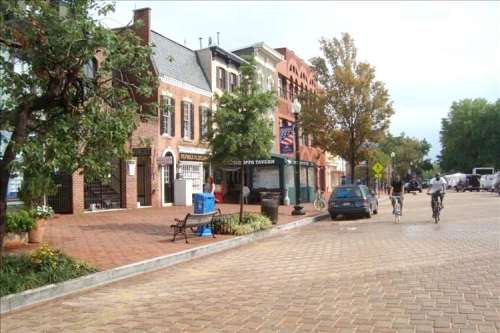 A charming street scene with brick buildings, trees, and cyclists on a cobblestone path.