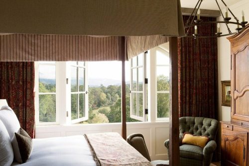Cozy bedroom with a four-poster bed, open windows, and a view of lush greenery outside. Elegant decor and warm lighting.
