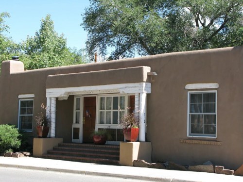 A single-story adobe-style house with a porch, surrounded by greenery and potted plants.