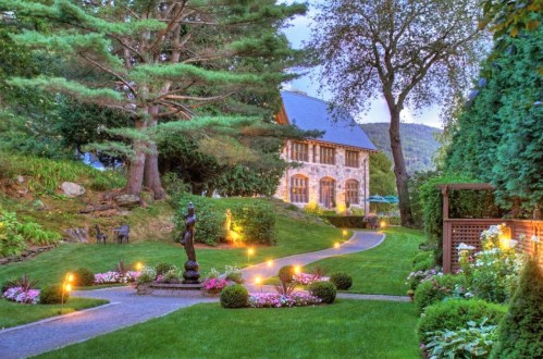 A beautifully landscaped garden with a stone house, pathway, and illuminated flower beds under a twilight sky.