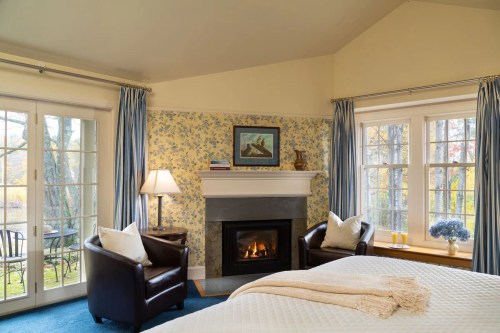 Cozy bedroom with floral wallpaper, a fireplace, and two armchairs by a window overlooking a serene outdoor view.