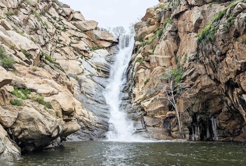 A waterfall cascades down rocky cliffs into a serene pool, surrounded by rugged terrain and sparse vegetation.