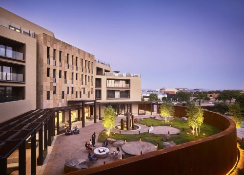 Modern hotel exterior with a landscaped courtyard, fire pit, and seating areas, set against a twilight sky.