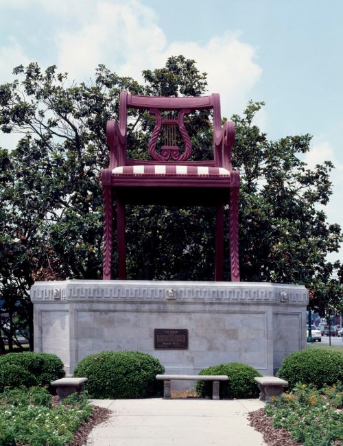 A large, decorative purple chair sculpture on a stone pedestal, surrounded by greenery and a blue sky.
