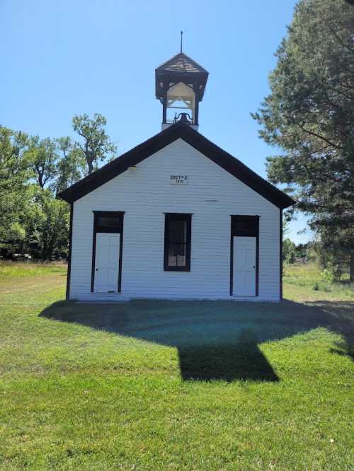 A small, white schoolhouse with a bell tower, surrounded by green grass and trees under a clear blue sky.