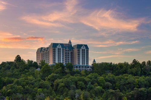 A large hotel building sits atop a green hillside under a colorful sunset sky.