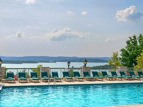 A serene poolside view with lounge chairs overlooking a lake and distant hills under a partly cloudy sky.