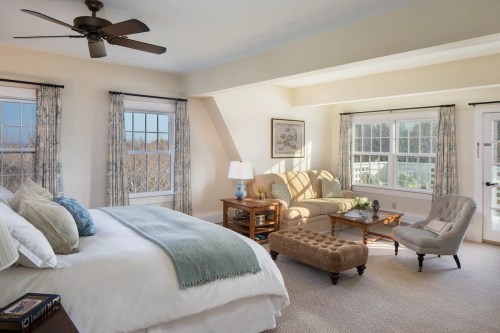 Bright, airy bedroom with a bed, seating area, and large windows, featuring light colors and cozy decor.