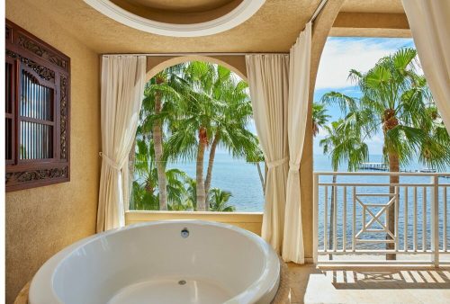 A luxurious bathroom with a large tub, overlooking palm trees and a serene ocean view through arched windows.
