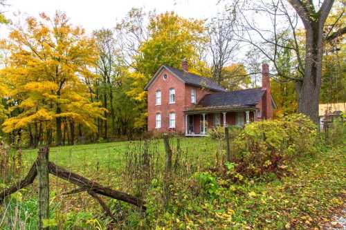 A brick house surrounded by colorful autumn trees and a grassy yard, with a wooden fence in the foreground.