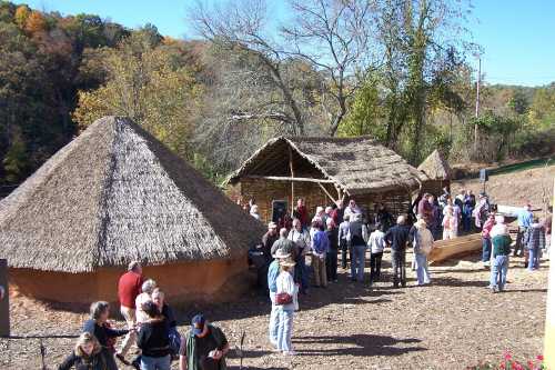 A crowd gathers near traditional thatched-roof structures in a wooded area, surrounded by autumn foliage.