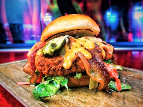 A close-up of a delicious burger with crispy chicken, bacon, cheese, pickles, and fresh greens on a wooden platter.
