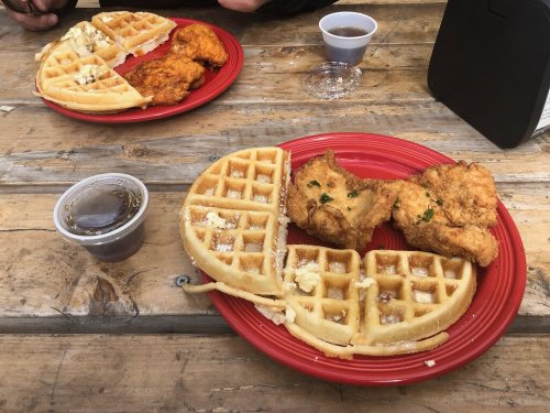 Two plates of chicken and waffles on a wooden table, with syrup in a cup and a drink in the background.