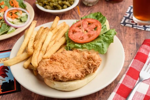 A plate featuring a fried chicken sandwich with lettuce, tomato, fries, and a side salad on a wooden table.