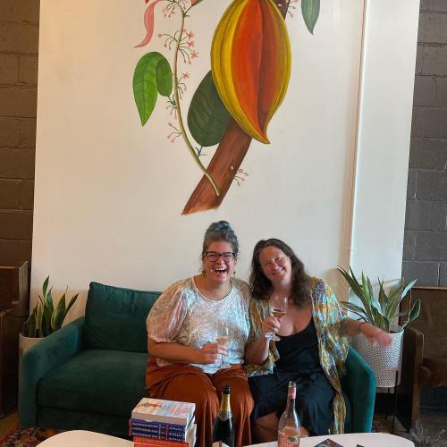 Two women sit on a green couch, smiling and holding glasses, with a colorful mural of a plant on the wall behind them.