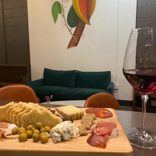 A wooden platter with cheese, meats, olives, and bread, alongside a glass of red wine, with a colorful mural in the background.