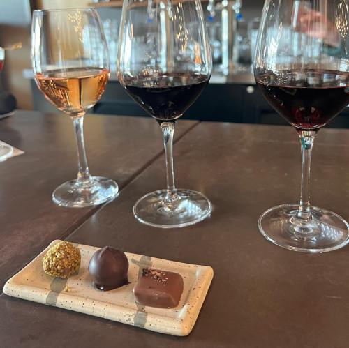 Three wine glasses filled with rosé and red wine, accompanied by a plate of two chocolates on a dark countertop.