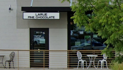 Exterior of LaRue Fine Chocolate shop, featuring a sign, outdoor seating, and greenery nearby.