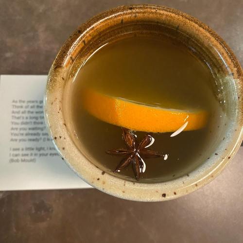 A warm drink in a ceramic cup with an orange slice and star anise, resting on a textured surface.