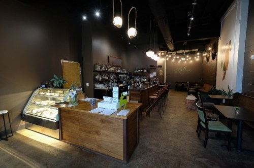 A cozy café interior featuring a wooden counter, bar seating, and a display case, with warm lighting and plants.