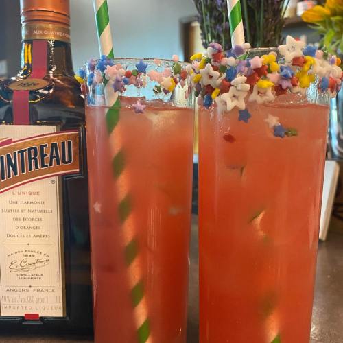 Two colorful cocktails with star-shaped sprinkles and striped straws, next to a bottle of Cointreau.