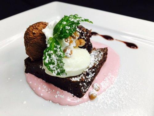 A decadent brownie topped with whipped cream, mint, chocolate mousse, and a drizzle of sauce on a white plate.