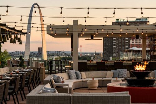 Rooftop lounge with string lights, seating area, and a view of the St. Louis Arch at sunset.