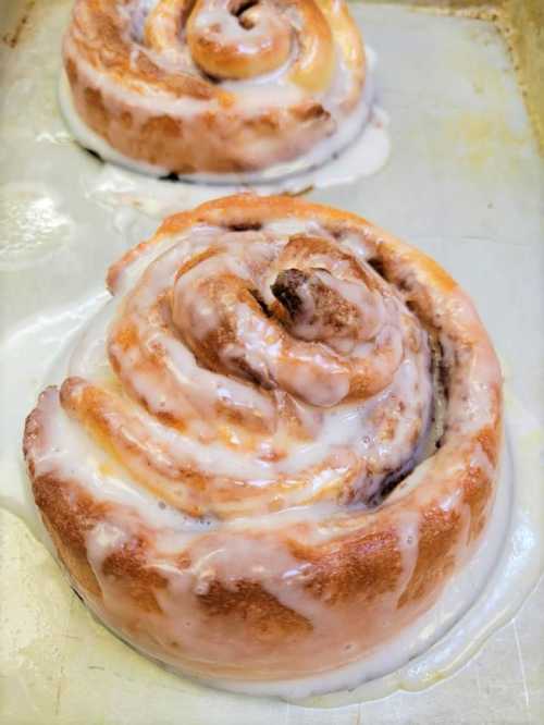 Two freshly baked cinnamon rolls drizzled with creamy icing, resting on a baking tray.