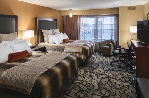 A cozy hotel room featuring two beds, a desk, a chair, and large windows with natural light.