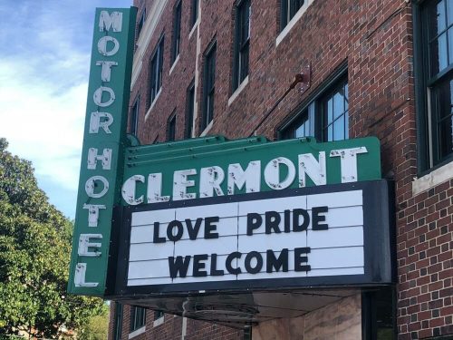 Sign outside the Clermont Motor Hotel displaying "LOVE PRIDE WELCOME" against a brick building backdrop.