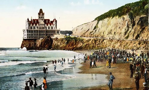 A vintage scene of a beach with people, waves, and a large castle-like building on a cliff in the background.