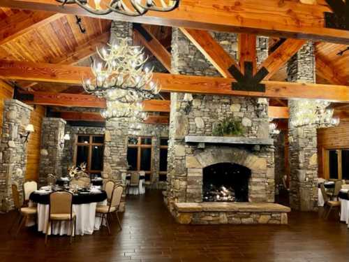 Cozy rustic dining area with stone walls, wooden beams, and elegant chandeliers, featuring a fireplace and round tables.