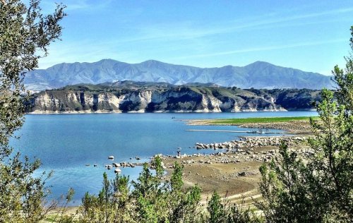 A serene lake surrounded by mountains, with clear blue water and rocky shores, framed by green foliage.