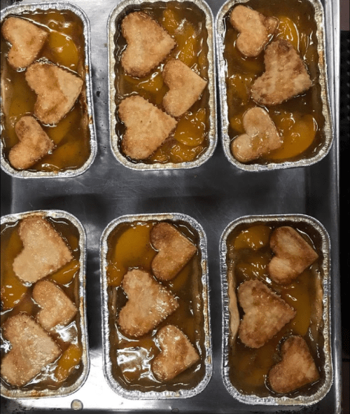 Six small aluminum trays filled with peach cobbler, topped with heart-shaped biscuit pieces.