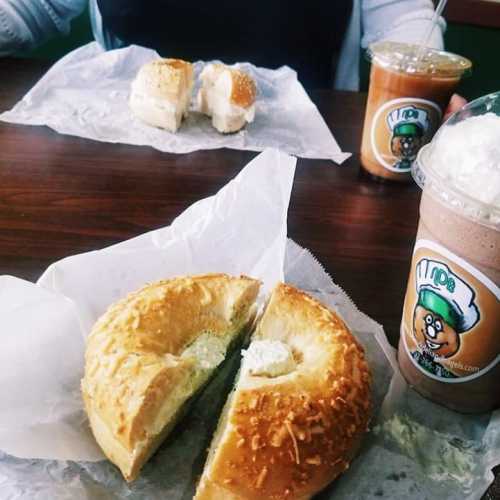 A table with two drinks and a bagel sandwich, one cut in half, on a paper wrapper.