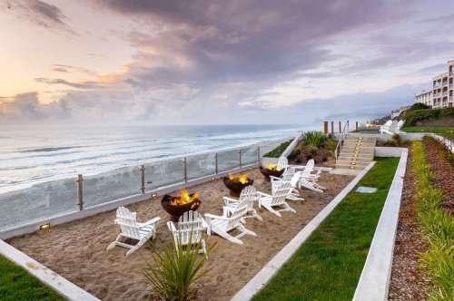 A serene beachside area with white chairs around fire pits, overlooking the ocean at sunset.