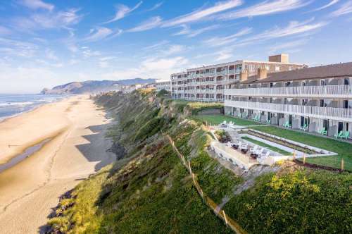 A coastal hotel overlooking a sandy beach, with green lawns and chairs, under a blue sky with wispy clouds.