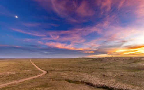 A winding dirt road stretches through a vast, open landscape under a colorful sunset sky.